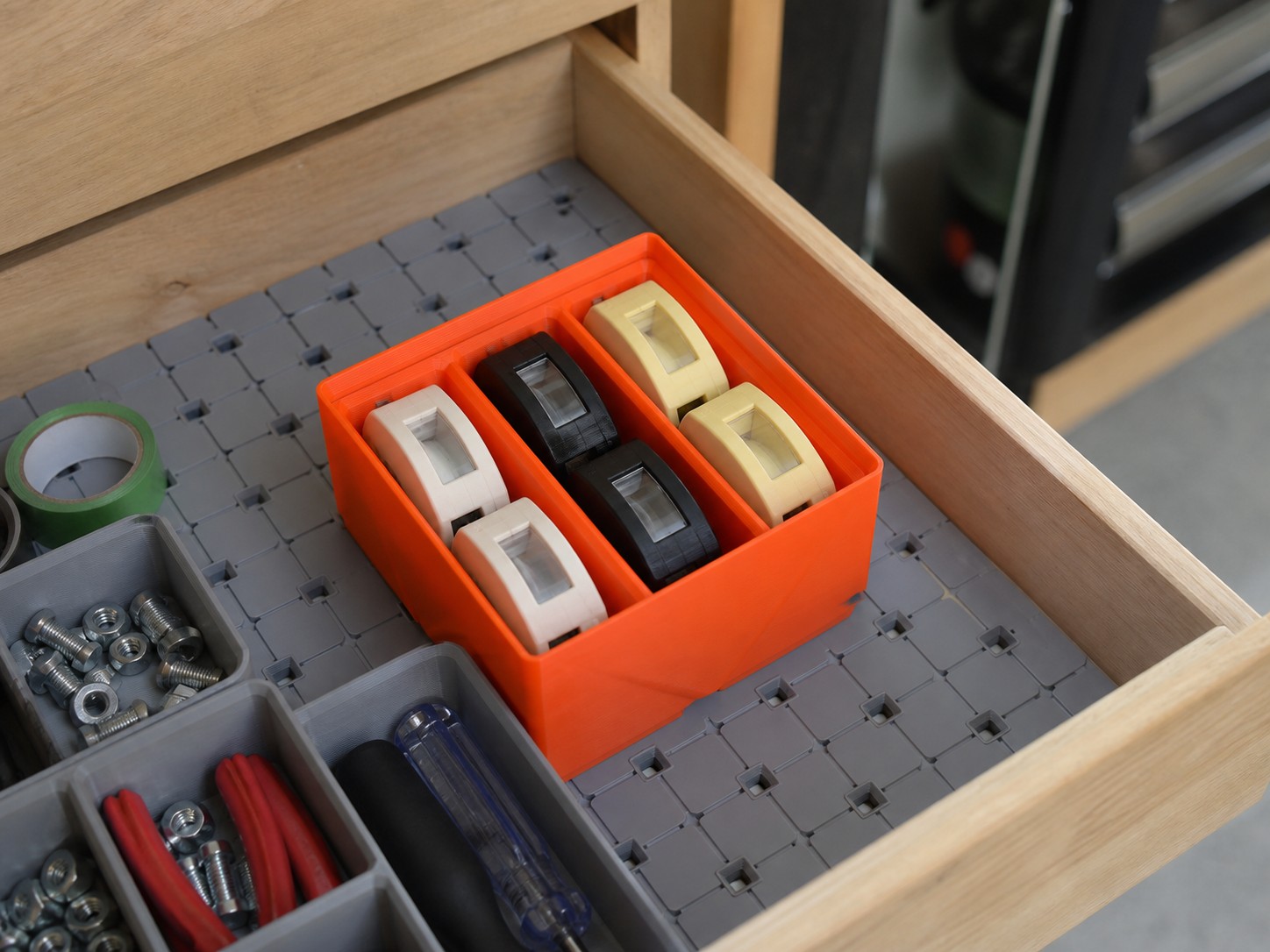 An orange printed bin holding label tape cartridges inside a workshop drawer lined with gray grid tiles, next to trays of bolts and pliers